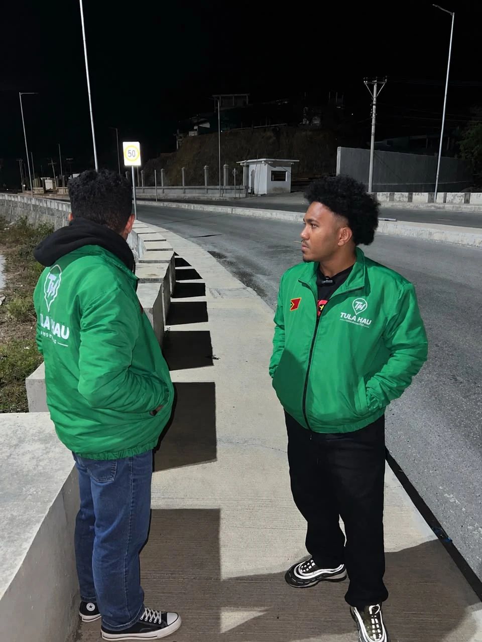 Two TULA-HAU drivers in green jackets talking at the roadside at night